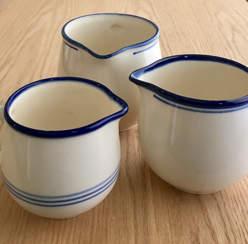 Pu&ograve; includere: Tre brocche di crema in ceramica bianca con bordo blu. Le brocche sono di dimensioni diverse e hanno una forma arrotondata.