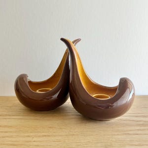 May include: Two brown ceramic candle holders with a glossy finish. The interior is a warm yellow-orange color. The candle holders have a unique, curved design, with a rounded base and a tall, arched top.