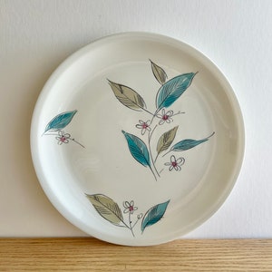 May include: A white ceramic plate with a floral design. The design features blue and green leaves and small white flowers with pink centers.