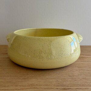 May include: A round, low-profile ceramic bowl in a pale yellow color with a speckled finish. The bowl features two small, integrated handles on opposite sides and a smooth, glossy surface that reflects light across its curved exterior.