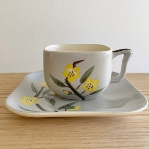 Puede incluir: Un juego de taza y platillo cuadrado gris con un diseño floral. La taza y el platillo tienen un fondo gris con flores amarillas y hojas verdes.