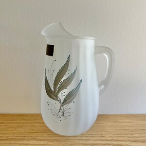 May include: A frosted glass pitcher with a floral design in shades of brown, green, and gold. The pitcher has a wide mouth and a handle.