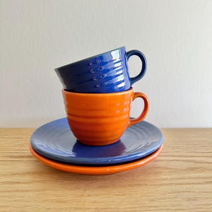 May include: A stack of two ceramic cups and two saucers. The bottom saucer is orange, topped with a blue saucer. An orange cup with horizontal ridges sits on the blue saucer, and a blue cup with horizontal ridges rests on top of it.