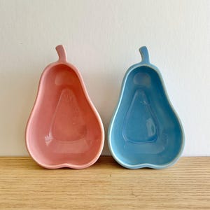 Könnte beinhalten: Zwei keramikförmige Schalen in Birnenform. Eine ist zartrosa, die andere hellblau. Beide haben ein Stieldetail oben und eine glatte, glänzende Oberfläche. Sie stehen nebeneinander auf einer Holzoberfläche vor weißem Hintergrund.