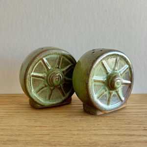 Puede incluir: Dos saleros y pimenteros de cerámica con forma de ruedas de carro. Los saleros y pimenteros están esmaltados en tonos verdes y marrones, con un aspecto rústico y texturizado. Cada uno tiene agujeros en la parte superior.