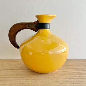 Puede incluir: Una jarra de cerámica amarilla brillante con un asa de madera y una banda negra alrededor del cuello. El asa está sujeta con herrajes de latón. La jarra tiene un cuerpo redondeado y un pico acampanado.