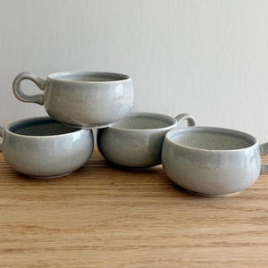 May include: A set of four light gray ceramic soup bowls with speckled textures and small, rounded side handles. Each bowl features a wide, open mouth and a smooth, curved body, arranged in a cluster to display their uniform shape and color.