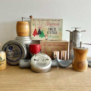 Può includere: Una collezione di utensili da cucina vintage, tra cui un macinapepe in legno, una scatola di tagliabiscotti e un tritatutto in metallo. È visibile anche una scatola di tagliabiscotti natalizi. Gli oggetti sono disposti su una superficie di legno.