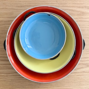 May include: A nested set of three colorful serving trays. The largest tray is red with black handles, the middle tray is yellow, and the smallest is blue. All trays have a silver trim.