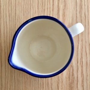 Pu&ograve; includere: Un'anfora di ceramica bianca con un bordo blu e un manico. L'anfora &egrave; vuota e ha un interno bianco liscio.