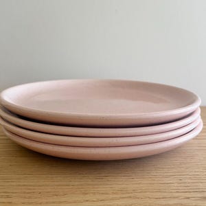 May include: A stack of four round ceramic plates in a soft pastel pink color. Each plate features a subtle speckled texture across the surface, a smooth glazed finish, and a slightly raised rim, creating a uniform and consistent appearance.