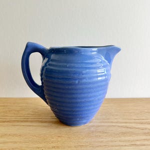 May include: A small, blue ceramic pitcher with a handle and a spout. The pitcher has a ribbed texture and is sitting on a wooden surface. The background is a plain white wall. This vintage pitcher is perfect for serving cream or milk.