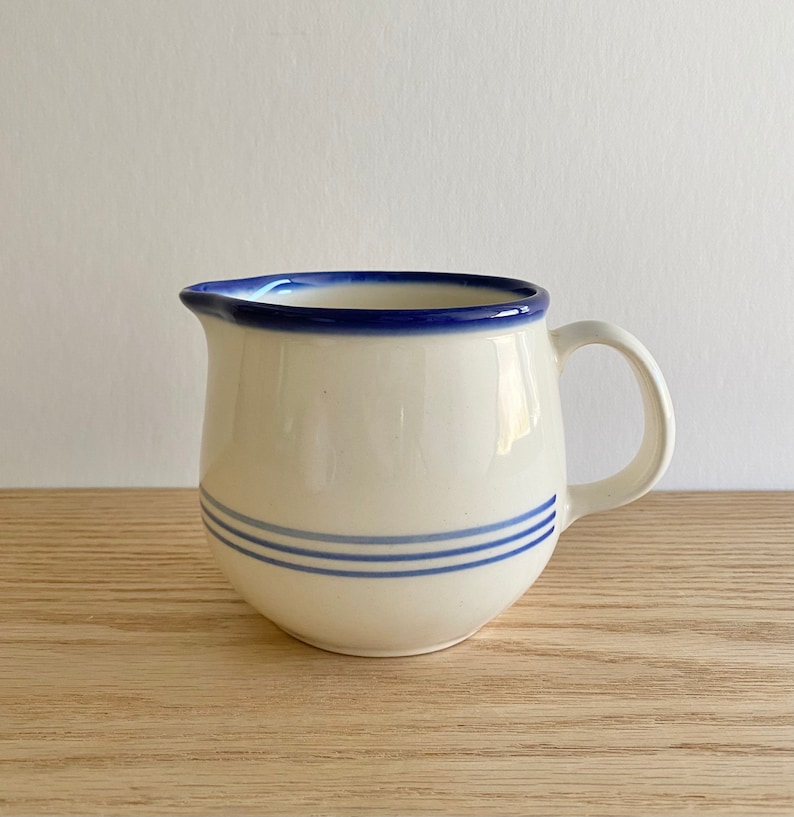 Pu&ograve; includere: Un brocca per la panna in ceramica bianca con un bordo blu e tre strisce blu intorno al centro. La brocca ha un manico ed &egrave; appoggiata su una superficie di legno.