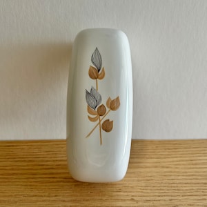 Puede incluir: Jarrón de cerámica blanco con un diseño floral estilizado en marrón y negro. El jarrón tiene una forma rectangular redondeada.