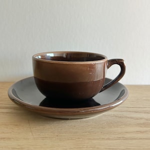 Puede incluir: Un juego de taza y platillo de cerámica marrón. La taza tiene un esmalte marrón con una banda marrón más oscura alrededor del medio. El platillo es negro.