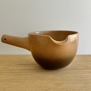 May include: A speckled, tan-colored ceramic bowl with a handle. The bowl has a rounded shape with a spout on one side. The handle extends horizontally from the bowl and has a small hole at the end. The bowl sits on a wooden surface.