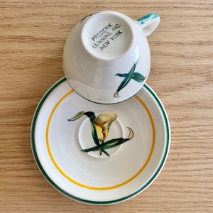 K&ouml;nnte beinhalten: Eine wei&szlig;e Teetasse und Untertasse mit gr&uuml;nem Rand und gelbem Band. Die Untertasse zeigt ein gemaltes Bild einer gelben Calla mit gr&uuml;nen Bl&auml;ttern. Die Teetasse tr&auml;gt den Schriftzug "FREDERIK LUNNING, INC. NEW YORK" auf der Unterseite.