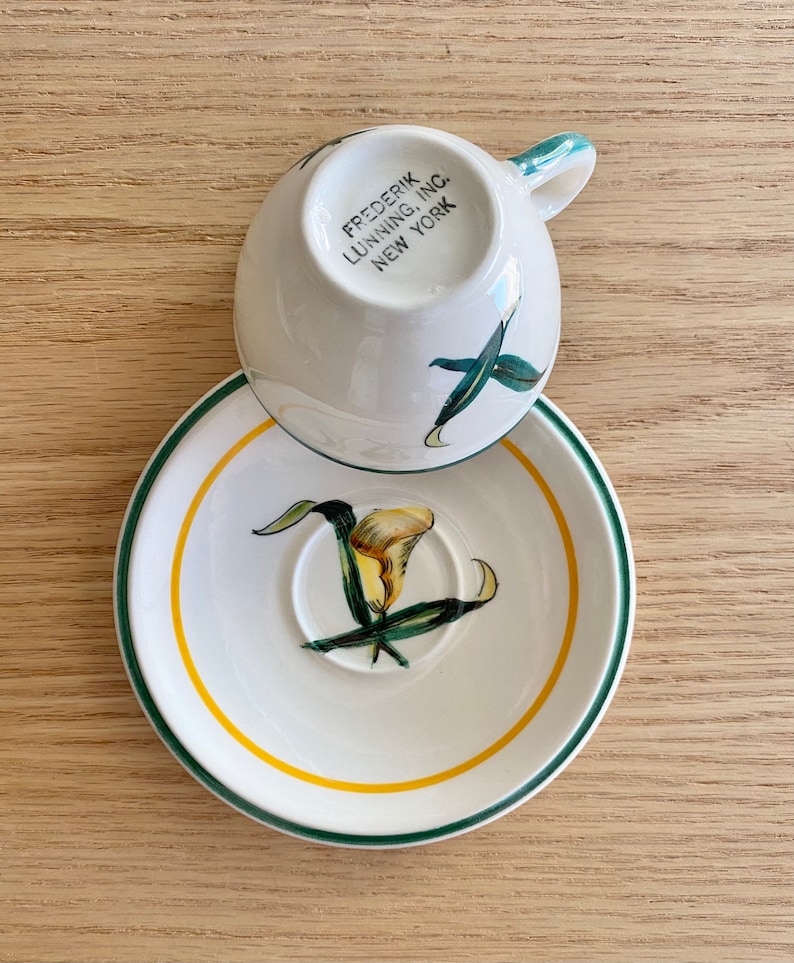 K&ouml;nnte beinhalten: Eine wei&szlig;e Teetasse und Untertasse mit gr&uuml;nem Rand und gelbem Band. Die Untertasse zeigt ein gemaltes Bild einer gelben Calla mit gr&uuml;nen Bl&auml;ttern. Die Teetasse tr&auml;gt den Schriftzug "FREDERIK LUNNING, INC. NEW YORK" auf der Unterseite.