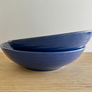 May include: Two blue ceramic bowls stacked on top of each other. The bowls have a wide, shallow design with a slightly textured surface. The bowls are a solid, deep blue color and appear to be handmade.