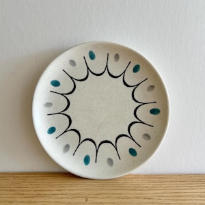 Puede incluir: Un plato redondo de cerámica blanquecina con una superficie moteada. El plato presenta un diseño negro festoneado alrededor del borde, con puntos turquesa y grises. El plato está sobre una superficie de madera.