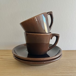 Puede incluir: Dos tazas de café de cerámica marrón apiladas una encima de la otra con un platillo marrón debajo. Las tazas tienen un acabado brillante.