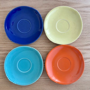 May include: Four ceramic saucers in different colors: blue, yellow, turquoise, and orange. Each saucer has a slightly raised rim and a small, circular indentation in the center.