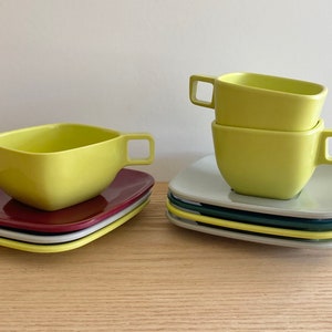 May include: A set of four yellow ceramic bowls, with two stacked on top of each other. The bowls are square shaped with rounded corners and have a slight lip around the top edge. The bowls are sitting on top of a stack of four saucers, two of which are grey, one is dark blue, and one is yellow. The saucers are also square shaped with rounded corners.