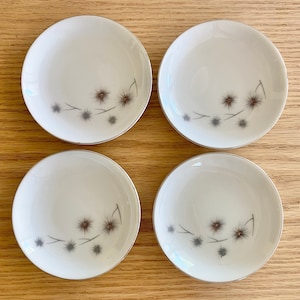 Peut inclure: Ensemble de quatre soucoupes en céramique blanche avec un bord argenté et un motif floral délicat dans des tons de gris.