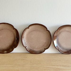 May include: Three ceramic plates with a distinctive scalloped edge. The plates feature a speckled, light brown centre and a darker brown rim. They are arranged on a wooden surface against a white background.