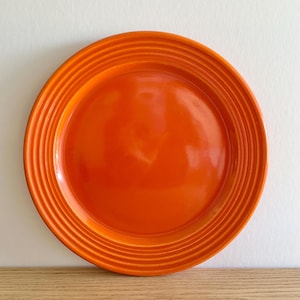 May include: A bright orange plate with a glossy finish. The plate has a series of concentric ridges around the rim, providing a textured detail. The plate is set against a neutral background, highlighting its bright colour and simple design.