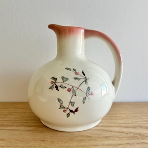 May include: A white ceramic pitcher with a round body and a handle. The rim and handle are a gradient of pink. Delicate floral branches with green leaves and red berries decorate the pitcher's surface. The pitcher sits on a wooden surface.