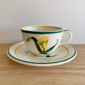 K&ouml;nnte beinhalten: Eine wei&szlig;e Teetasse und Untertasse aus Keramik mit einem gr&uuml;nen und gelben Blumendesign. Die Tasse hat einen Henkel und die Untertasse einen gr&uuml;nen Rand.