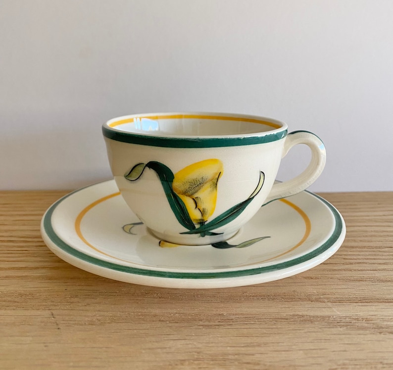 K&ouml;nnte beinhalten: Eine wei&szlig;e Teetasse und Untertasse aus Keramik mit einem gr&uuml;nen und gelben Blumendesign. Die Tasse hat einen Henkel und die Untertasse einen gr&uuml;nen Rand.