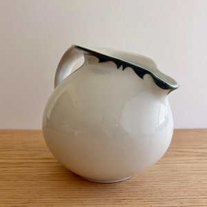 May include: A round, white ceramic pitcher with a smooth, glossy finish. The vessel features a curved handle on the left side and a distinct, dark green painted rim that follows the contour of the pouring spout, creating a sharp contrast.
