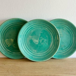Peut inclure: Trois assiettes en céramique turquoise avec des motifs de cercles concentriques. Les assiettes sont disposées sur une surface en bois sur fond blanc. Les assiettes ont une finition brillante et une texture légèrement irrégulière.