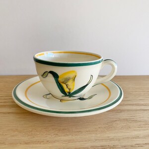 K&ouml;nnte beinhalten: Eine wei&szlig;e Teetasse und Untertasse aus Keramik mit einem gr&uuml;nen und gelben Blumendesign. Die Tasse hat einen Henkel und die Untertasse einen gr&uuml;nen Rand.