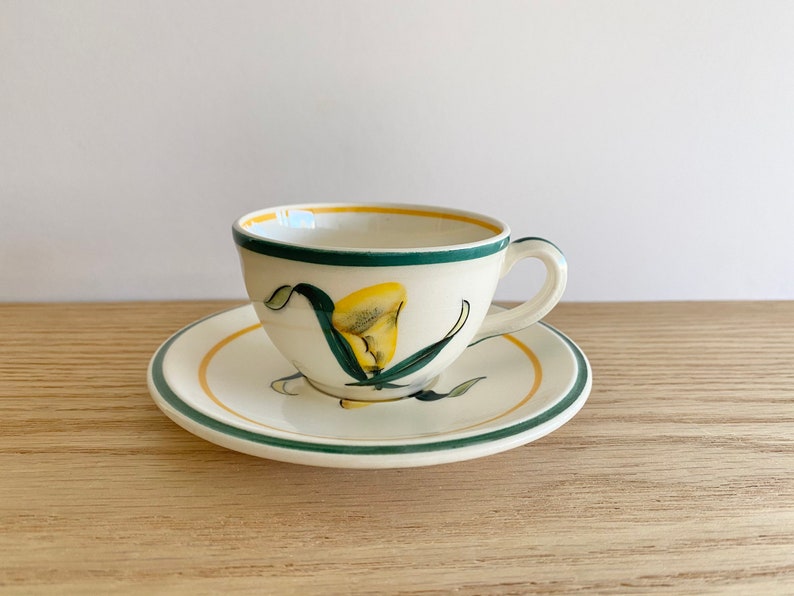 K&ouml;nnte beinhalten: Eine wei&szlig;e Teetasse und Untertasse aus Keramik mit einem gr&uuml;nen und gelben Blumendesign. Die Tasse hat einen Henkel und die Untertasse einen gr&uuml;nen Rand.