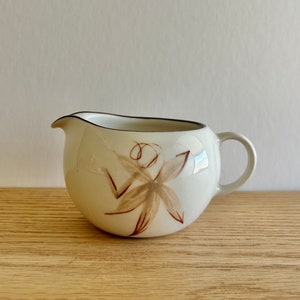 May include: A white ceramic creamer with a brown floral design. The creamer has a rounded body and a small handle.