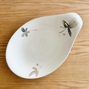 May include: White, teardrop-shaped serving dish with floral designs in black, gray, and tan. The dish has a smooth surface and a slightly raised edge. The floral designs are delicate and add a touch of elegance to the dish.
