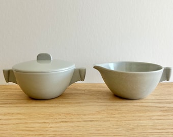 May include: A light gray ceramic sugar bowl with a lid and a matching creamer. The sugar bowl has a rounded shape with two small handles. The creamer has a spout and a single handle. Both pieces are on a wooden surface.