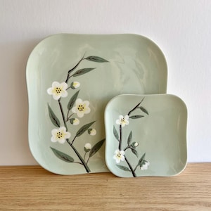Puede incluir: Dos platos cuadrados de cerámica verde claro. El plato más grande es rectangular con esquinas redondeadas, y el más pequeño es cuadrado. Ambos platos presentan un diseño floral con flores blancas, centros amarillos y hojas verdes.