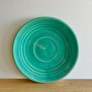 May include: A turquoise ceramic saucer with concentric rings. The saucer has a central indentation and a slightly raised rim. The color is a vibrant teal, and the surface has a glossy finish. The saucer is likely for a teacup or coffee cup.