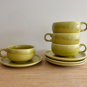 May include: A set of four olive green ceramic soup bowls with matching saucers. One bowl sits on a saucer to the left, while three bowls are stacked vertically on top of three saucers to the right. Each bowl features a single rounded handle.