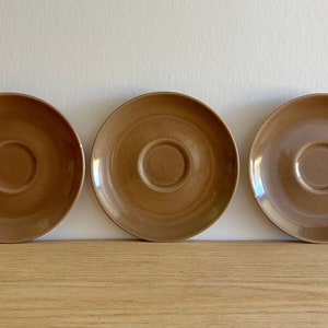 May include: Three brown ceramic saucers with a slightly raised center. The saucers are arranged in a row on a wooden surface.