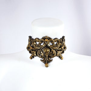 May include: A gold-toned metal choker necklace with an intricate, ornate design featuring a bird skull in the center. The necklace is made of a thick, durable metal and has a vintage, gothic aesthetic.