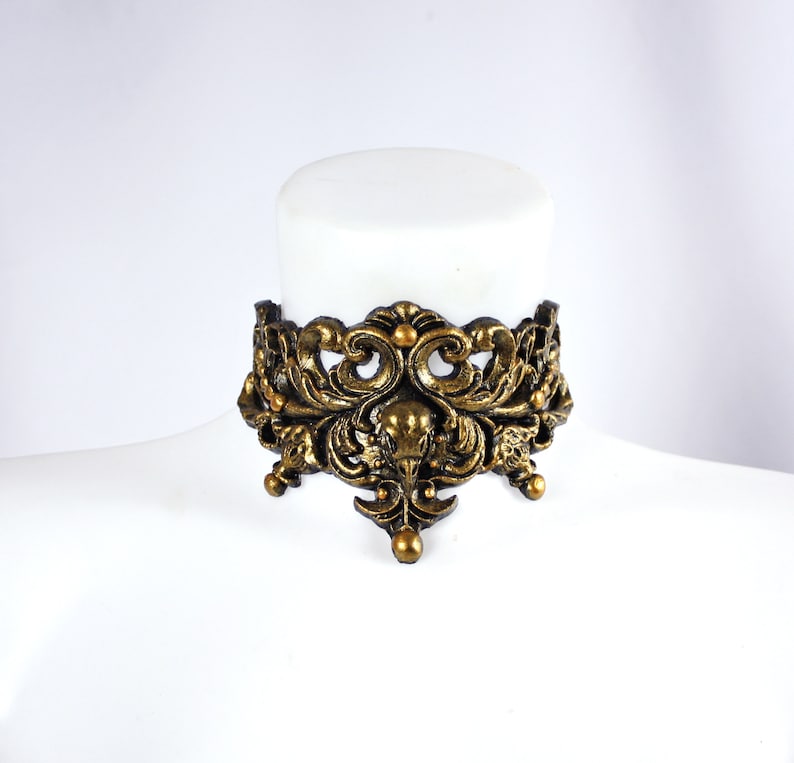 May include: A gold-toned metal choker necklace with an intricate, ornate design featuring a bird skull in the center. The necklace is made of a thick, durable metal and has a vintage, gothic aesthetic.