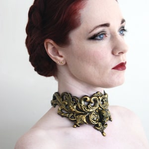May include: A woman with long, dark red hair styled in a braid, wearing a gold and black ornate choker necklace.