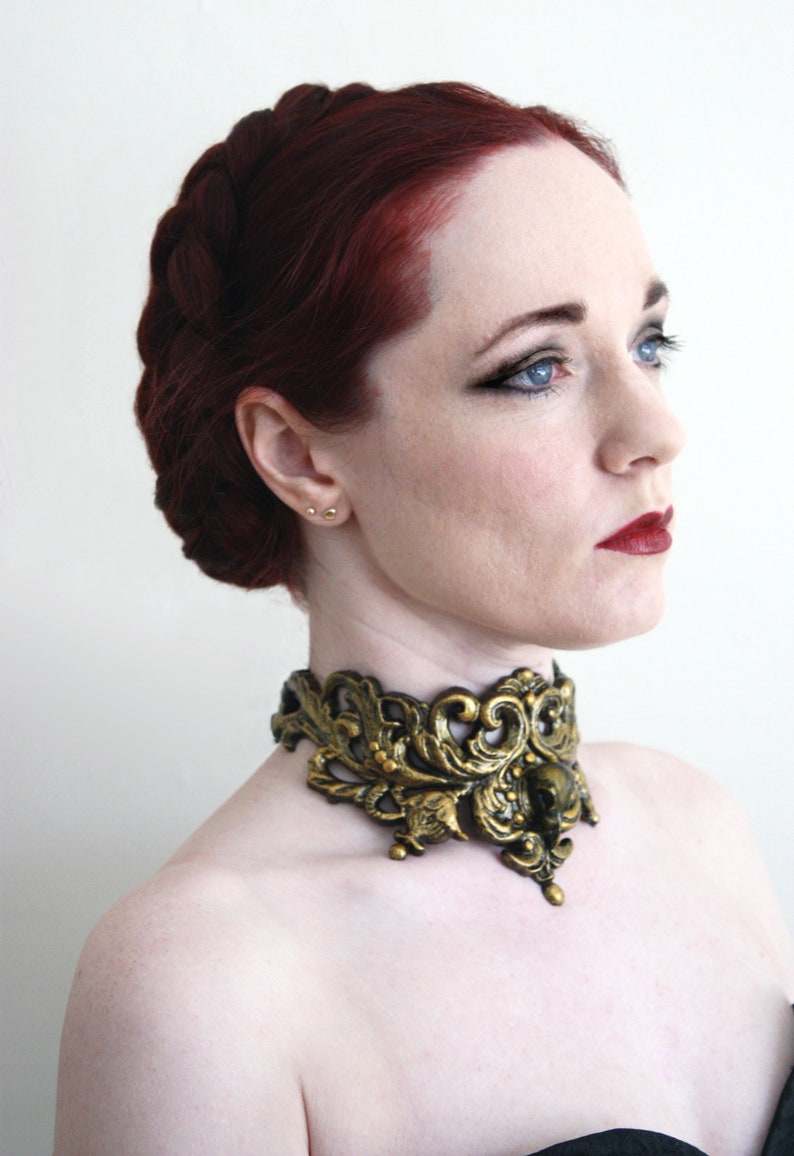 May include: A woman with long, dark red hair styled in a braid, wearing a gold and black ornate choker necklace.