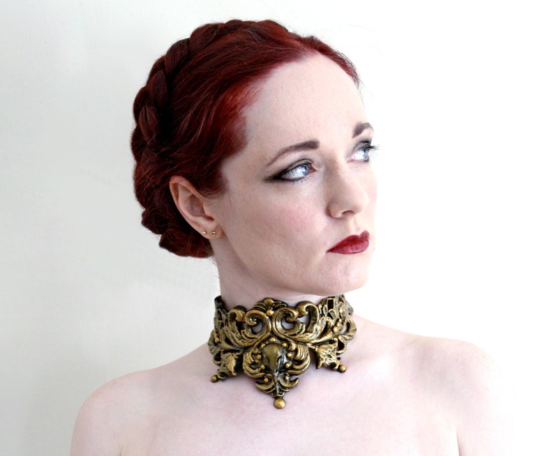 May include: A woman with red hair wearing a gold, ornate, decorative choker necklace.