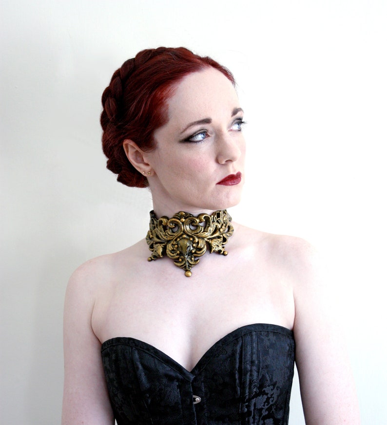 May include: A woman with red hair wearing a black strapless top and a gold ornate choker necklace.